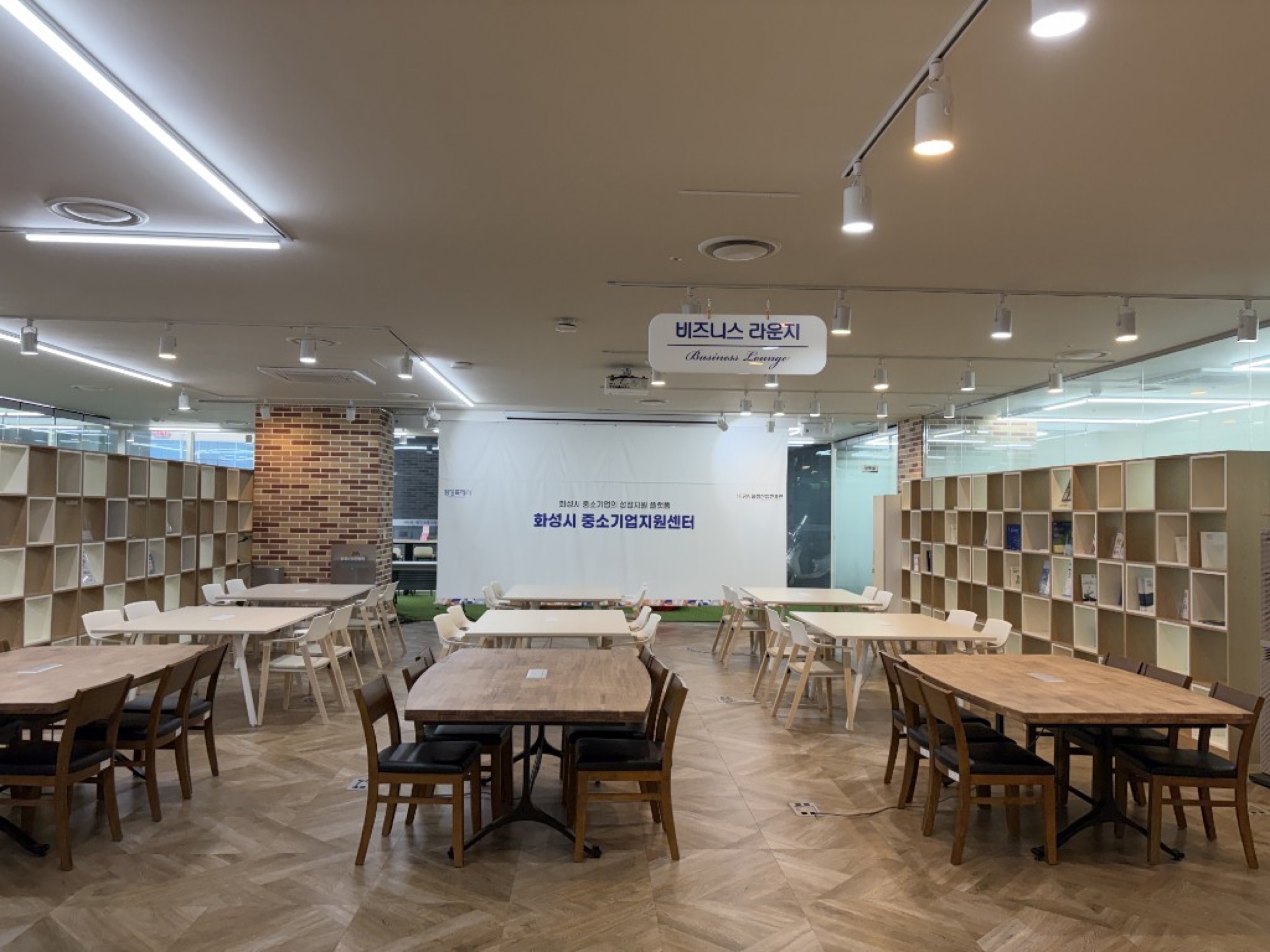 헤링본 마루와 나무 테이블, 양쪽 책장이 있는 화성특례시 중소기업지원센터 비즈니스 라운지. 천장에 비즈니스 라운지 안내판이 걸려 있다.